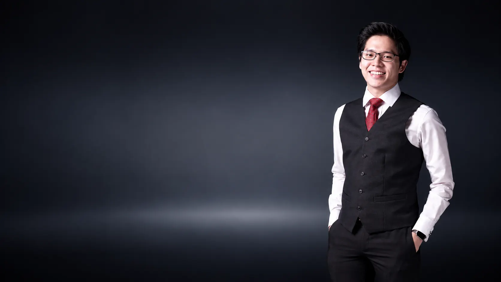 Dr. Jovian Tan smiling, standing with hands in pockets in a white shirt, charcoal waistcoat, red patterned tie and black-rimmed glasses, against a dark studio background